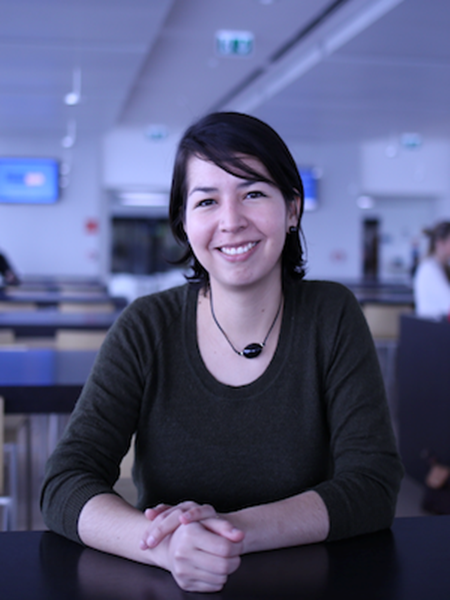 Reina is a woman from Venezuela. She has black short hair parted to to left side, with a subtle fringe. She is wearing a black t shirt with mid-length sleeves. She has a necklace on with black thread and black solid stone pendant. She is sitting against a table with her hand folded, and smiling while directly looking at the camera. Her background is blurred but it looks like she is in a university class room.