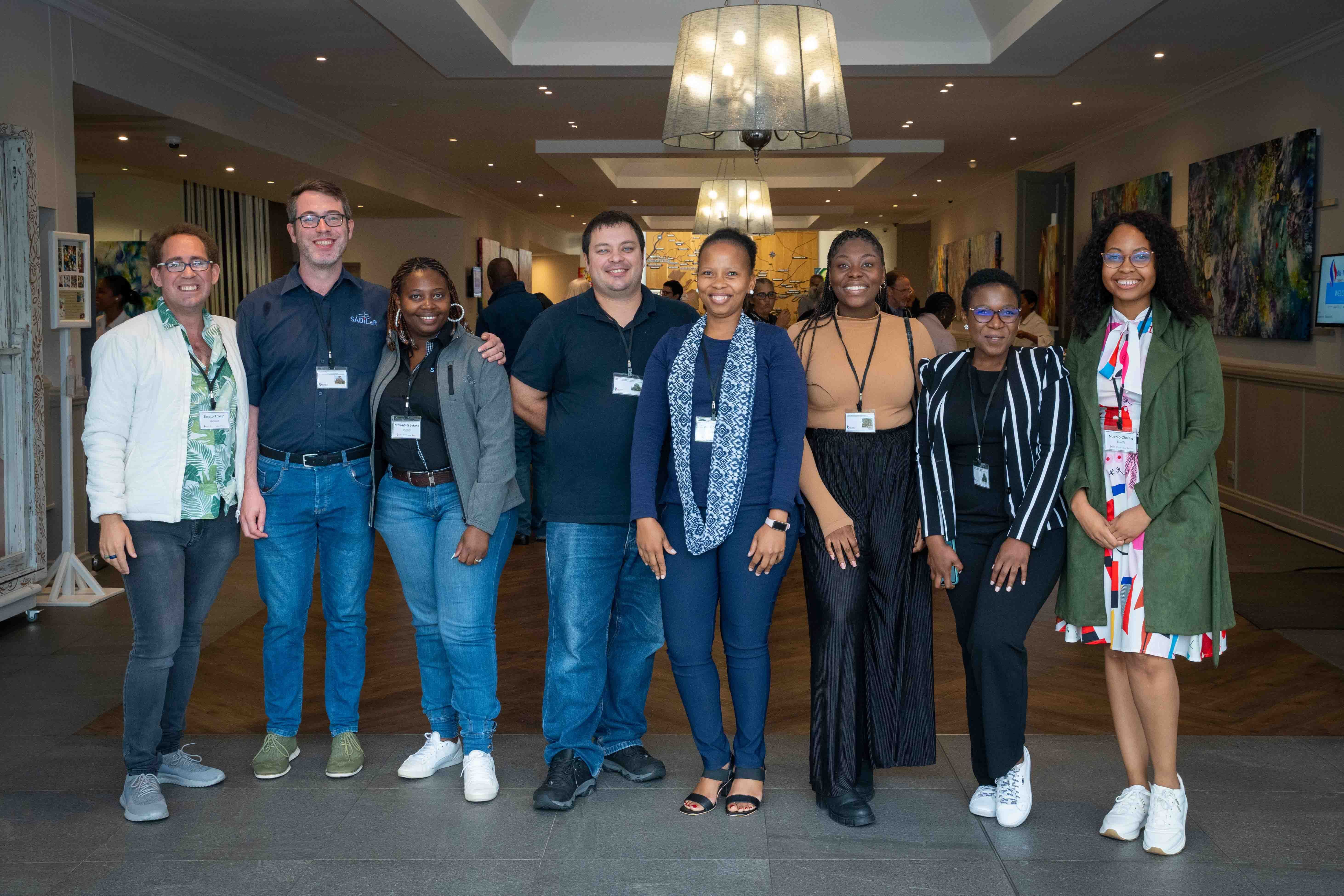 eight people posing for the camera inside a fancy looking building, with lots of led lights on the ceiling. A very well framed photo. They seem to be at a conference venue lobby. They all wear credentials and their clothes are colourful and semi-formal. They are all smiling and look very happy. Three men and four women. Two women are wearing white sneakers